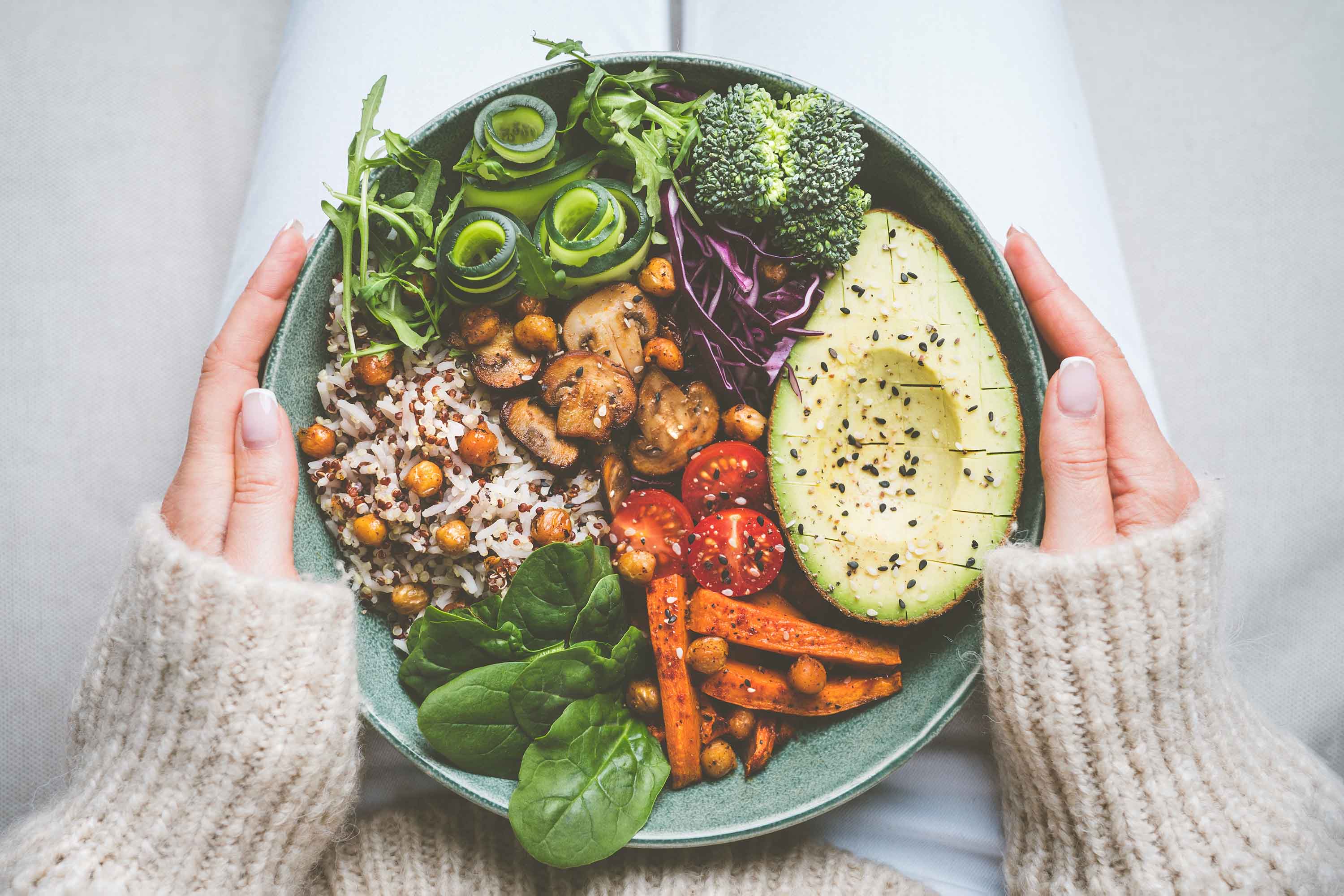  A person holding a bowl of plant-based food with avocado, mushrooms, sweet potato, broccoli, cucumber, and spinach.